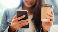 Asian Woman Happy And Typing Messages On Smartphone In Cafe. Smiling Young Woman Sitting At Table With Hot Cup Of Coffee And Using Mobile Phone