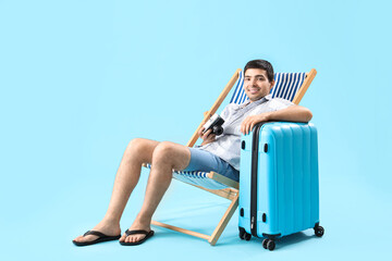 Poster - Young man with photo camera and suitcase sitting in deck chair on blue background