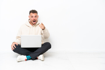 Wall Mural - Young caucasian man with a laptop sitting on the floor isolated on white background surprised and pointing front