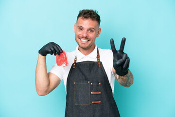 Wall Mural - Butcher caucasian man wearing an apron and serving fresh cut meat isolated on blue background smiling and showing victory sign