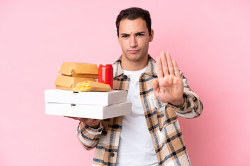 Wall Mural - Young caucasian man holding fast food isolated on pink background making stop gesture