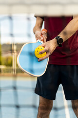 Wall Mural - Front view of a senior man posing with a pickleball racket and a ball in sportswear. The man is seen through the net of a sports court. Pickle ball concept.