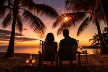 Christmas On The Tropical Coast, New Year Mood. Rear View Of Young Happy Couple In Love Sitting On The Chairs On The Beach And Watching Fireworks.