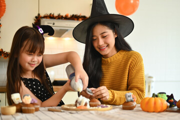 Happy mother with little daughter preparing delicious sweet dishes for Halloween party in kitchen