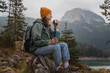 Young cheerful woman traveler with a backpack and thermos on the shore of a lake in the mountains. Solo travel concept