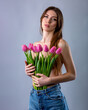 Middle-length studio portrait of a beautiful sexy slim young caucasian brown-haired girl topless in blue jeans holding bouquet of pink tulips