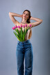 Middle-length studio portrait of a beautiful sexy slim young caucasian brown-haired girl topless in blue jeans holding bouquet of pink tulips in her jeans