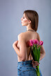 Studio portrait of a beautiful sexy slim young caucasian brown-haired girl topless with silver chain necklace holding bouquet of pink tulips behind her back with one hand and her breast with another