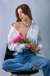 Studio portrait of a beautiful sexy slim young caucasian brown-haired girl in white shirt and blue jeans with silver chain necklace sitting and holding bouquet of pink tulips in her hands