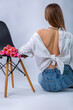 Studio portrait of a beautiful sexy slim young caucasian brown-haired girl in white shirt and blue jeans with silver chain necklace sitting near a bouquet of pink tulips shot from her back