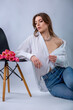 Studio portrait of a beautiful sexy slim young caucasian brown-haired girl in white shirt and blue jeans with silver chain necklace sitting near a bouquet of pink tulips