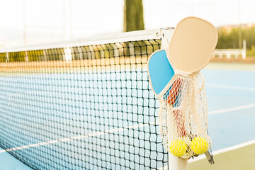 a mesh bag with a pair of pickleball rackets and two balls hooked to a net post on a sports court. p