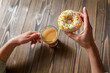 Female hands holding cup of coffee and donut in a white glaze with yellow confectionery sprinkles on a wooden background.