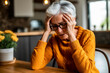 © Nedrofly - A depressed older woman sitting at a table with her hands on her head