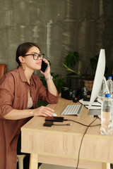 Aufkleber - Young busy woman speaking on smartphone and reviewing new shots on computer screen after photo session while sitting by workplace