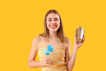 Wall Mural - Beautiful happy young woman with glass of tasty cocktail and shaker on yellow background