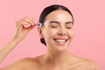 Wall Mural - Beautiful young woman applying serum onto her face on pink background