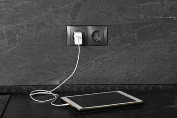 Wall Mural - Tablet charging from electric socket on dark grey table indoors