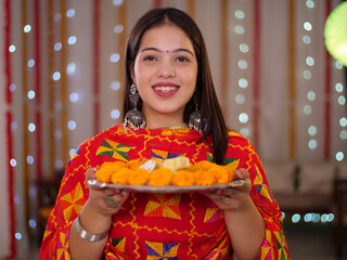 An attractive Indian woman happily smiling and offering sweets - posing for the camera - Diwali festival. Beautiful young female offering barfi with a wide smile on her face - decorative festive ba...