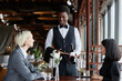 © Seventyfour - Portrait of Black young man as server taking orders from couple in luxury restaurant and presenting bottle of red wine, copy space