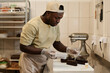 Side view portrait of young Black man as male baker decorating chocolate pastries in bakery, copy space