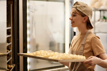 Side view portrait of smiling young woman putting fresh raw breads in freezer at bakery kitchen, copy space