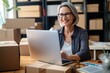 © Stavros - Online store seller during an online conversation with a buyer. Smiley middle aged Caucasian woman sits in front of laptop monitor in a warehouse of products during online video call with a customer.