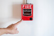 © Summer Paradive - Emergency fire warning system pull station. Hand pointing at red fire alarm mounted on white wall in public building