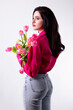 Studio portrait of a beautiful sexy slim young caucasian brunette girl in pink shirt and jeans holding bouquet of pink tulips shot from her back and looking dreamily to the left