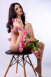 Studio portrait of a naked beautiful sexy slim young caucasian brunette girl covered with pink butterflies holding a bouquet of pink tulips sitting on chair attractively posing looking in camera