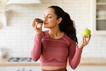 Fitness Woman Choosing Donut Instead Of Healthy Apple In Kitchen