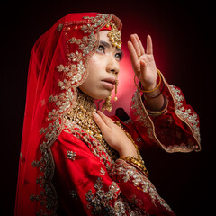 Wall Mural - Close up portrait of a beautiful Asian Muslim lady in a hijab wearing a gorgeous Bollywood or Indian themed red traditional wedding dress isolated on dark background