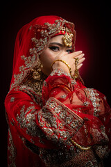 Wall Mural - Close up portrait of a beautiful Asian Muslim lady in a hijab wearing a gorgeous Bollywood or Indian themed red traditional wedding dress isolated on dark background