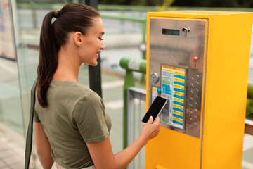 Wall Mural - Young woman standing on bus stop paying ticket with smartphone