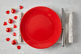 Fototapeta  - Red ceramic plate with cutlery on a white wooden tray surrounded by red Christmas balls and hearts on a light concrete table. Template for displaying New Year's food