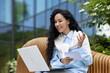 © Liubomir - Young beautiful woman sits on a bench outside an office building, uses a laptop to watch a webinar course video, a student writes data in a notebook, video call with a teacher, distance learning.