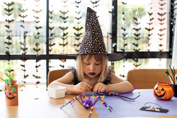 Wall Mural - A cute girl in a witch costume makes a funny spider for Halloween with her own hands. Children's crafts for Halloween