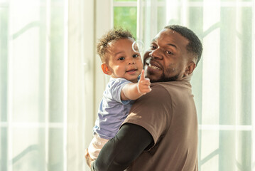 Wall Mural - Happy african black parents dad father throw baby son on piggyback and neck riding teasing fun near window. Black baby son and daddy enjoy teasing kiss cheek and neck riding at window light