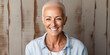 A bald senior caucasian woman with a shaved head with short hair smiling, wearing white top with blue jean jacket standing in front of wooden panel background