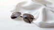 © พงศ์พล วันดี - A pair of aviator sunglasses on a white silk scarf. This image represents a fashionable and sophisticated style. The sunglasses have a gold frame and dark lenses, giving a cool and confident look.