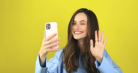 Wall Mural - Happy brunette woman having mobile video call, wave hands and say hi, hello isolated on yellow background. Nice to see you. Happy emotions. Beautiful young girl speaking video call online smartphone.