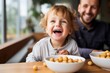 Happy toddler child boy eating a small bowl full of food. Parent is looking after him on the background