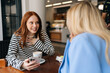 © dikushin - Side view of two happy beautiful women friends sitting in cafe indoors, looking at phone and talking, laughing. Cheerful pretty females girlfriends using social media on smartphone in coffee shop.