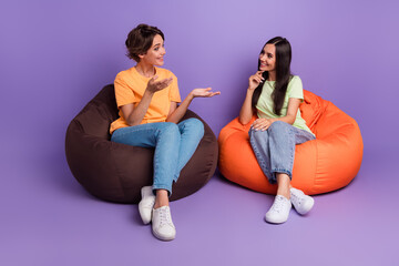 Wall Mural - Photo of cheerful positive ladies wear t-shirts sitting bean bags talking isolated violet color background