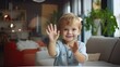 © lililia - a cheerful child sitting on a modern living room sofa, waving with enthusiasm and connecting with friends and family through a webcam.