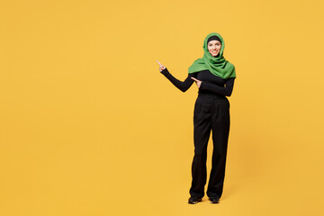 Wall Mural - Full body young smiling happy arabian asian muslim woman wear green hijab abaya black clothes point index finger aside on area isolated on plain yellow background. People uae islam religious concept.