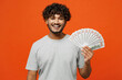© ViDi Studio - Young smiling satisfied happy Indian man he wears t-shirt casual clothes hold in hand fan of cash money in dollar banknotes isolated on orange red color background studio portrait. Lifestyle concept.
