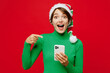 © ViDi Studio - Young fun woman wear warm cozy green turtleneck Santa hat posing hold use point finger on mobile cell phone isolated on plain red background. Happy New Year 2024 celebration Christmas holiday concept.