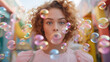 Young Pretty Girl with Curly Hair in Pink Dress Blowing Bubble, Ball of Chewing Gum. Brightly Colorful Building on Background. National Chewing Bubble Gum Day.  High quality photo