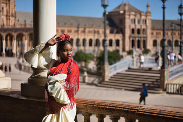 Wall Mural - Young black and South American woman in a beige gypsy flamenco suit and red shawl, dancing in a beautiful square in the city of Seville in Spain. Concept dance, folklore, flamenco, art.